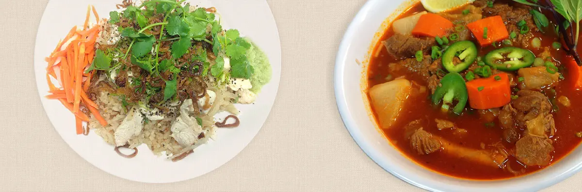 A Vietnamese rice entree next to a bowl of Vietnamese curry.  The rice is topped with carrots, fried shallots, chicken, cilantro, egg, and a green sauce.  The curry is red and contains beef, potatoes, carrots, jalapeno peppers, basil, green onions, and a lime.